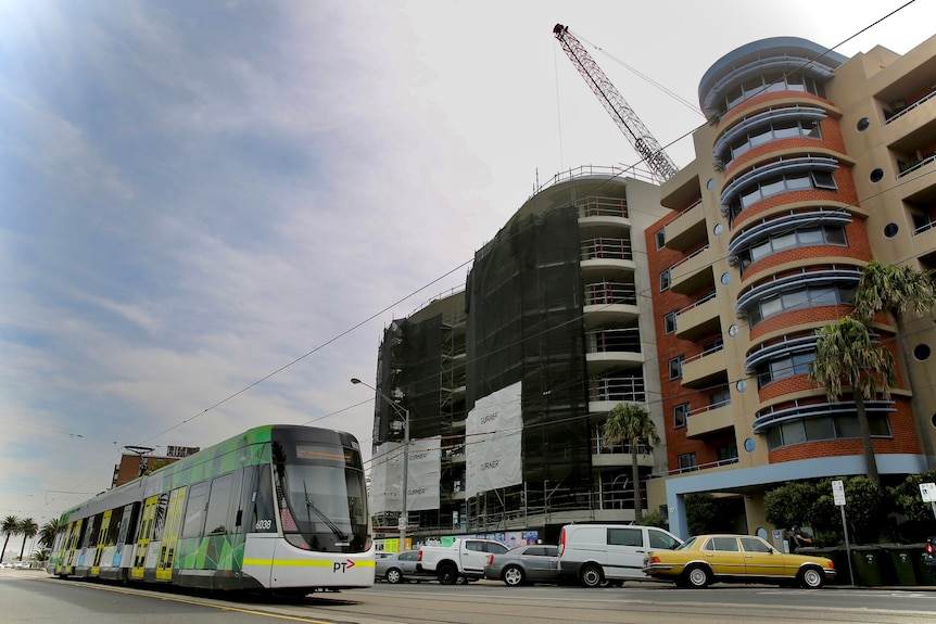 A tram moves past a construction site for an apartment building on a street in St Kilda on a cloudy day.