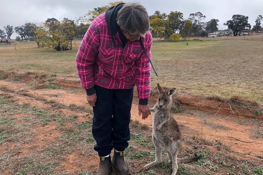 A woman in a pink flannelette shirt, black trousers, and boots bends down and holds the hand of a joey.