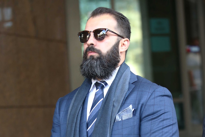 A man wearing sunglasses, with a beard and wearing a suit leaves a court building.