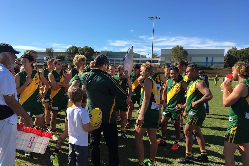 Coach with board in hand addresses football team in green/yellow uniforms, water runner and boy watch on