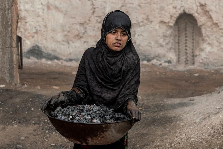 une fillette de 9 ans porte une casserole pleine de débris dans une briqueterie, les mains couvertes de poussière noire
