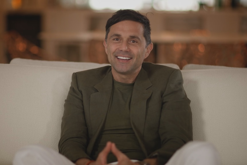 Steven Nasteski sitting on a white cream couch with his hands together in his lap wearing a khaki suit jacket smiling