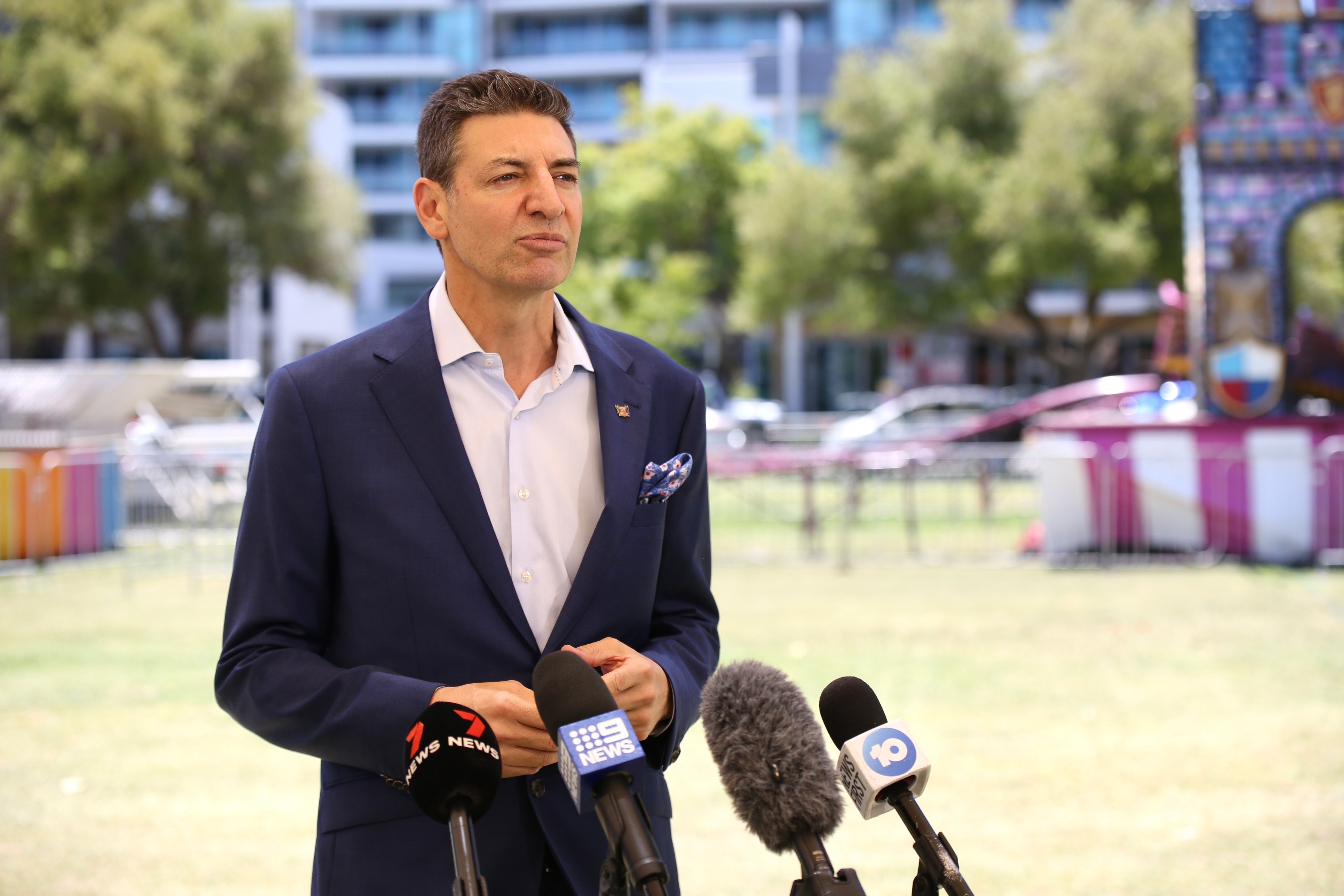 Basil Zempilas speaking into microphones outdoors on a sunny day, wearing a dark suit and white shirt.