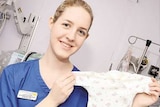 A young woman wearing a blue nurse's uniform smiles while holding up a babygrow