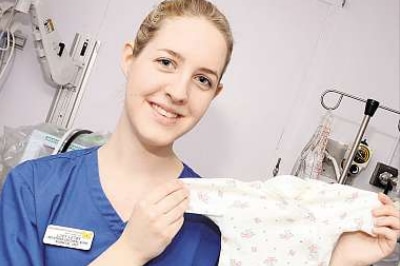 A young woman wearing a blue nurse's uniform smiles while holding up a babygrow