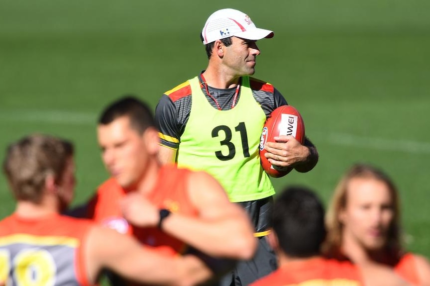 A man with a white hat and a fluorescent T-shirt holds a red AFL ball.