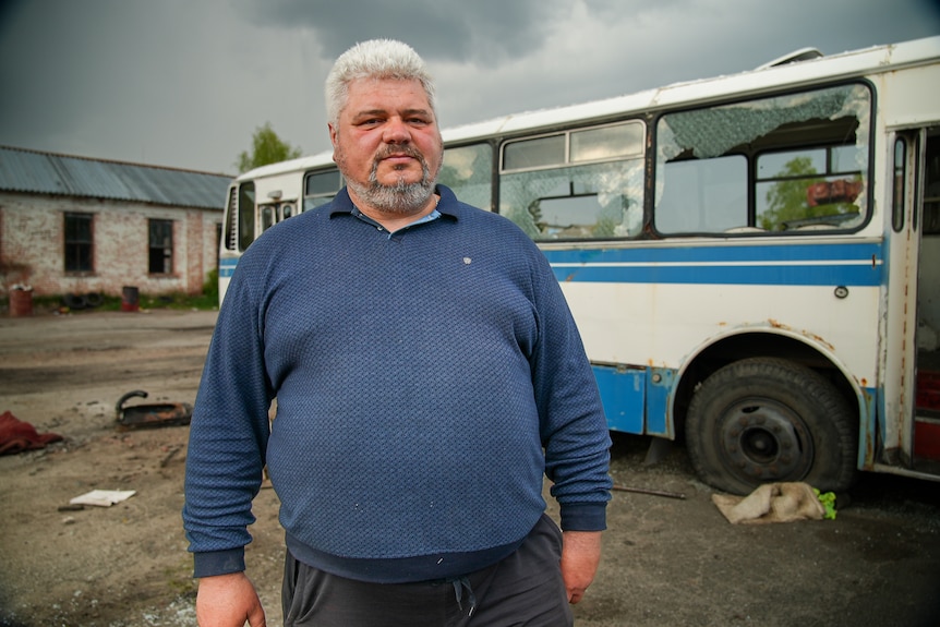 Un hombre canoso con un polo azul se para al lado de un autobús volado en pedazos. 