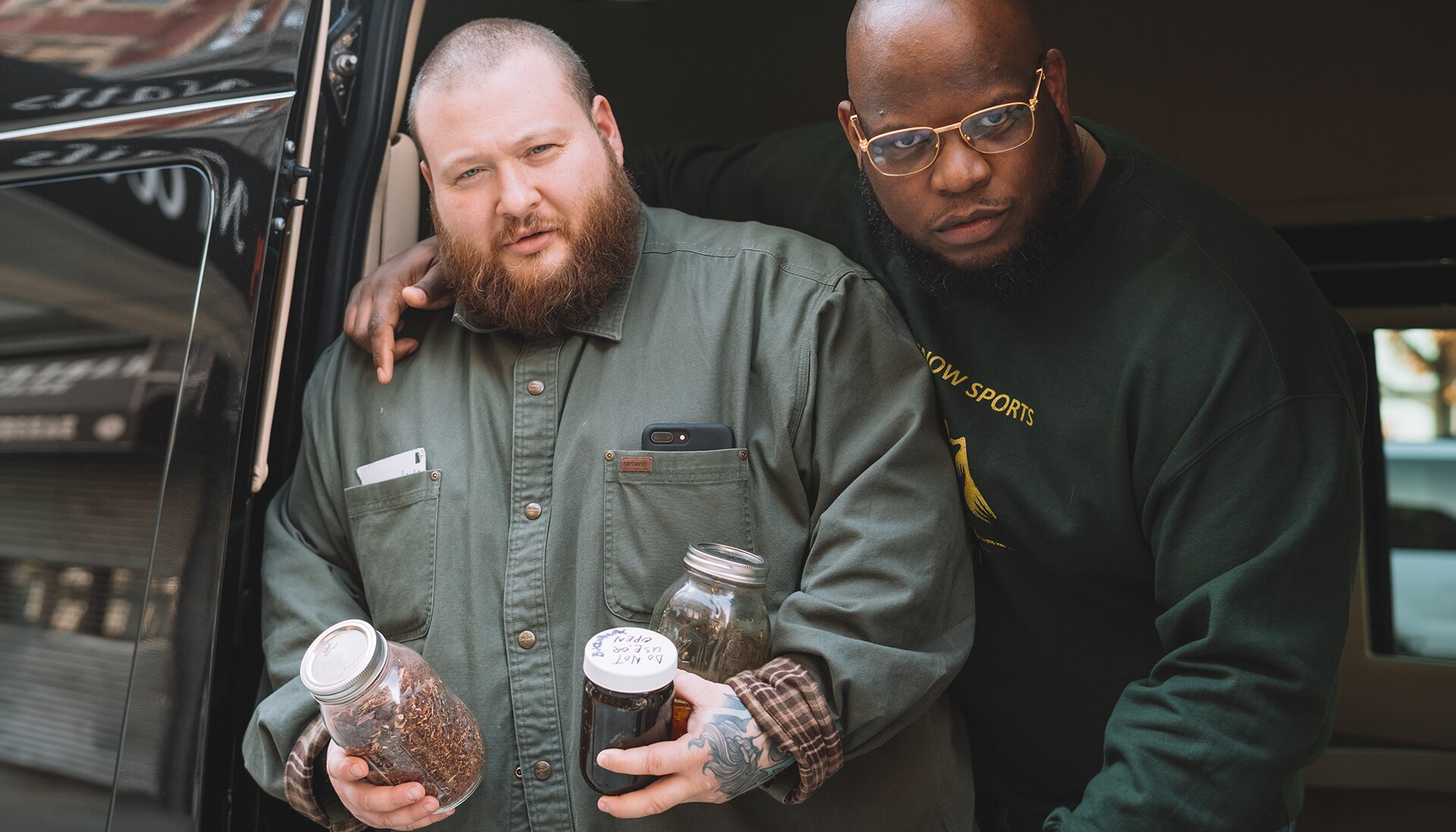 Two men stand looking at the camera, one holding jars of food