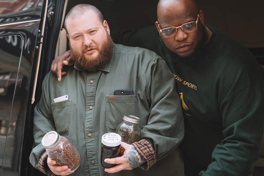 Two men stand looking at the camera, one holding jars of food