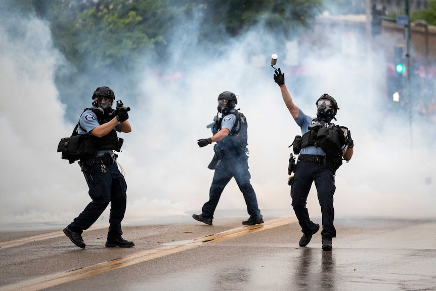 Police officers wearing gas masks walk in smoke as one throws a tear gas cannister.