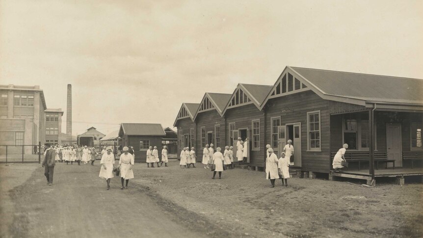 Sepia photo of female factory workers in white coats walking on a gravel road and standing next to wooden houses