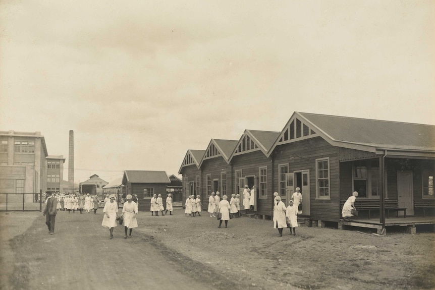 Sepia photo of female factory workers in white coats walking on a gravel road and standing next to wooden houses