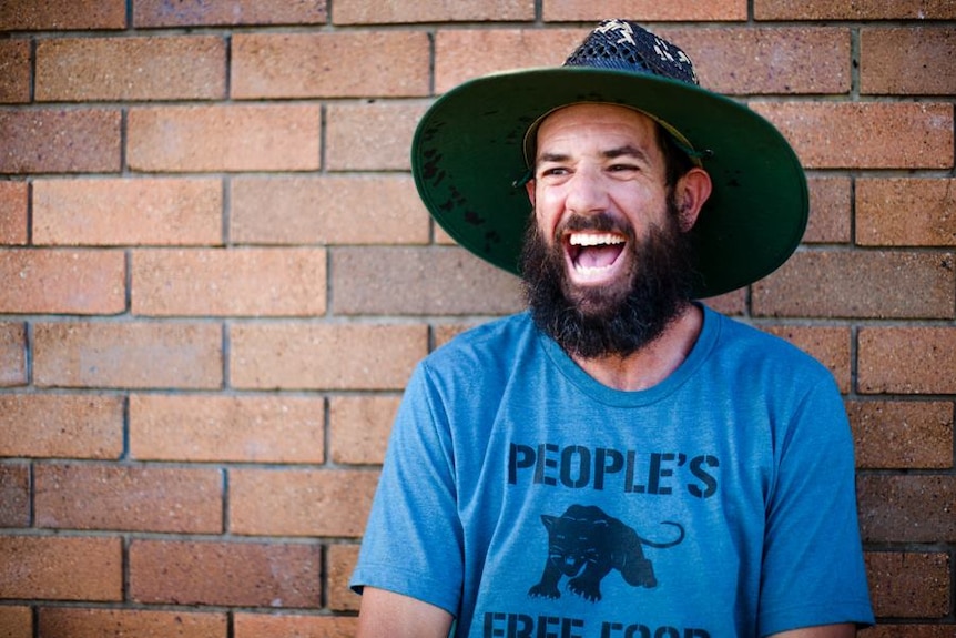 A man in a broad-brimmed hat laughs as he sits in front of a brick wall.