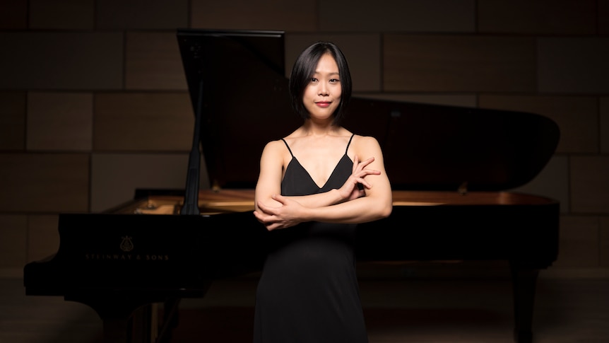 Yeol Eum Son stands with her arms crossed in front of a grand piano.
