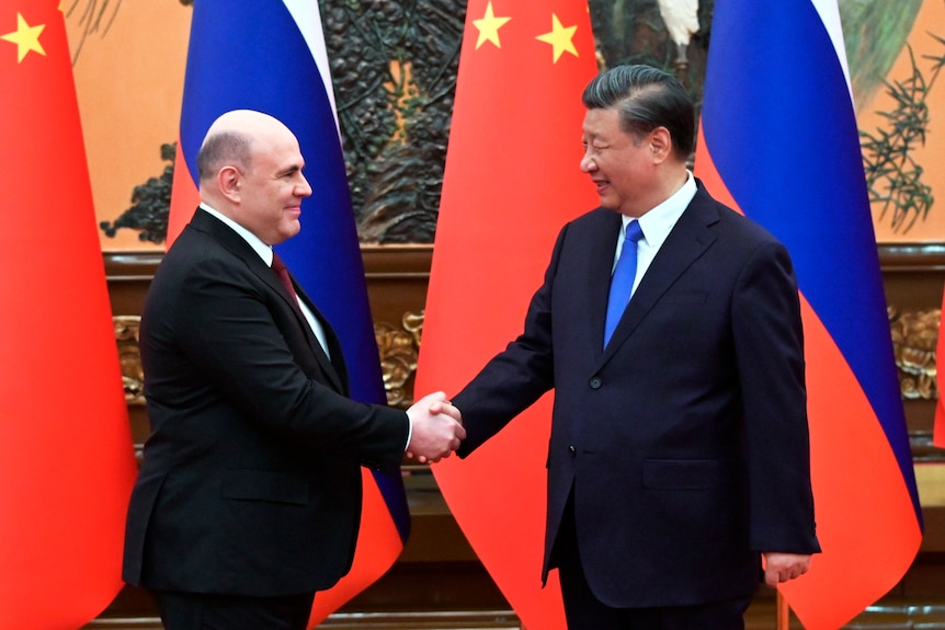 Mikhail Mishustin Xi Jinping shake hands while smiling and standing near Russian and Chinese flags.
