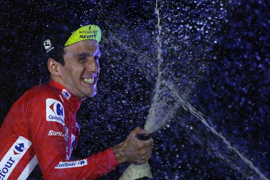 Simon Yates wears a red jersey and sprays champagne