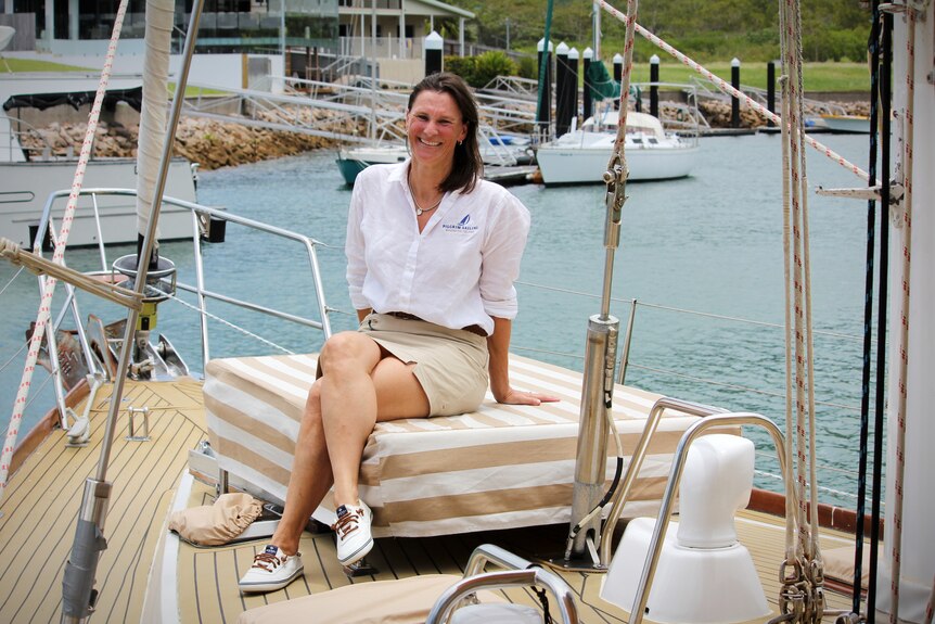 A smiling woman wearing a 'Pilgrim Sailing' shirt sits on the deck of a yacht docked in a marina. 