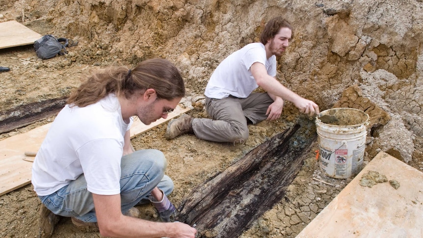 Two men sit scraping dust off a thin, wooden coffin in a trench.
