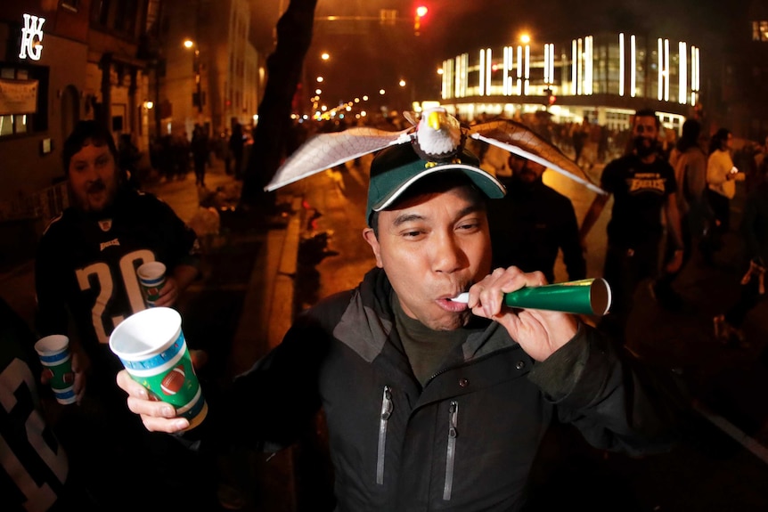 A Philadelphia Eagles fan wears an eagle on his baseball cap and blows a party horn in the street