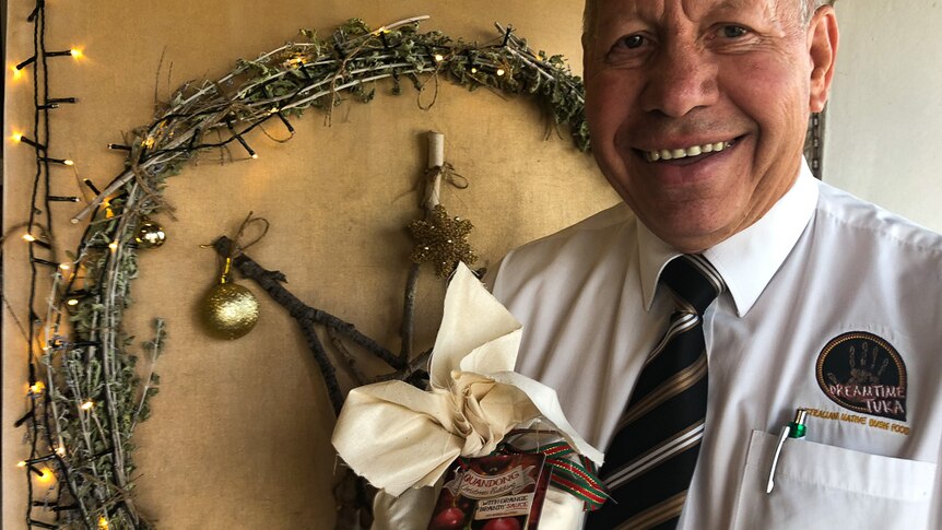 A man in a shirt and tie smiles in front of a Christmas wreath adorned with lights while holding a packaged Christmas pudding.