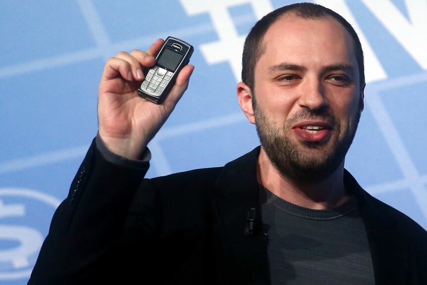 Jan Koum holds up a mobile phone in front of a blue-and-white background.