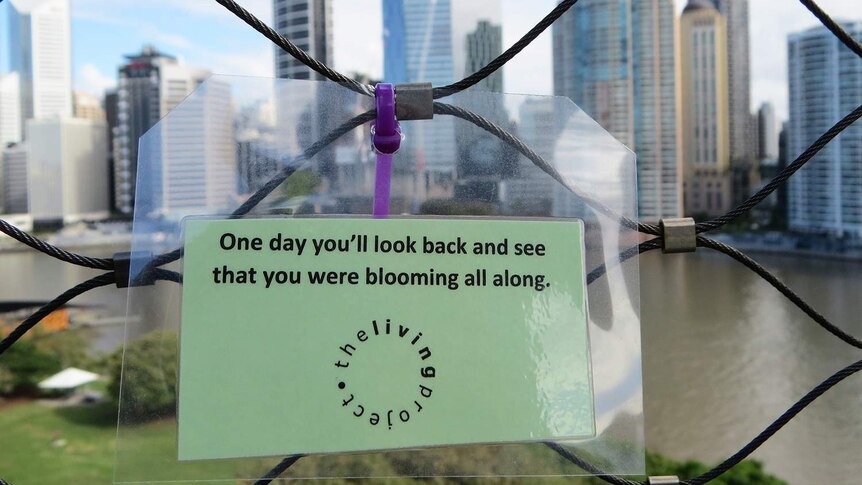 Laminated sign 'One day you'll look back and see that you were blooming all along' zip-tied on a bridge.