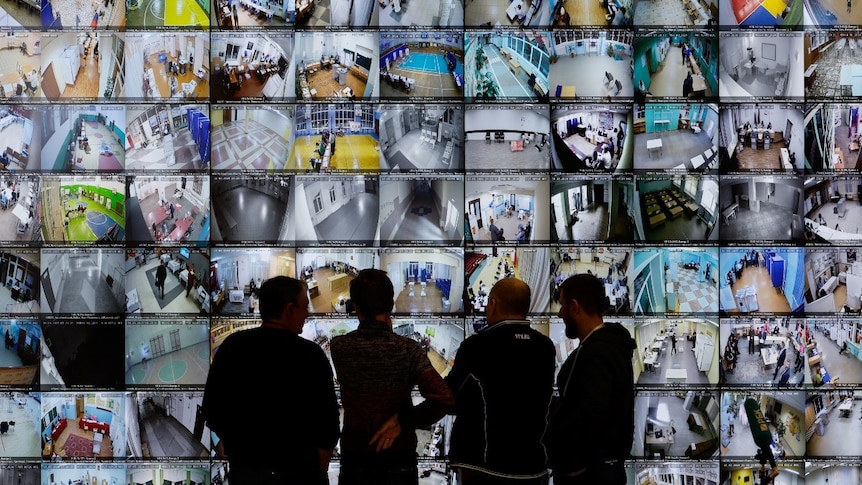 Four people stand in front of a large wall of computer screens, showing CCTV footage on them