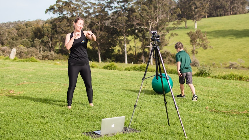 A woman in black lycra boxing  in green field in front of a camera on tripod, as a boy plays with a fitness ball.