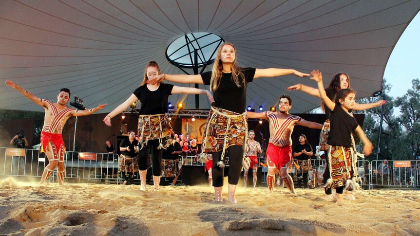 Young Aboriginal men and women dance on a sandy floor.