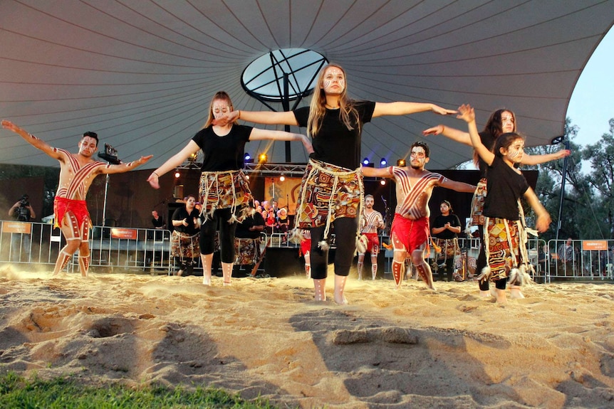 Young Aboriginal men and women dance on a sandy floor.