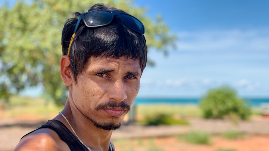 A man with dark hair  and a small face tattoo stares moodily at the camera, with blue sky and trees in the background.