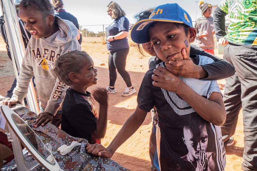 A group of children play with their faces in front of a mirror with red dirt behind.