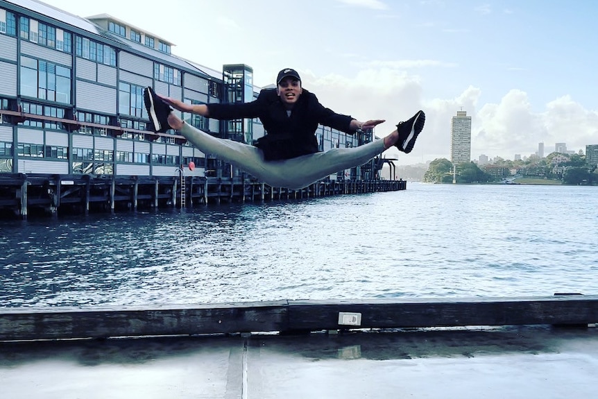 A young man does the splits in the air in front of the water