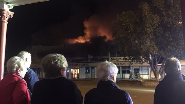 View of the back of five people watching flames and smoke pour out of the top of a building across the road from them