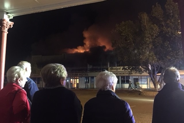 View of the back of five people watching flames and smoke pour out of the top of a building across the road from them