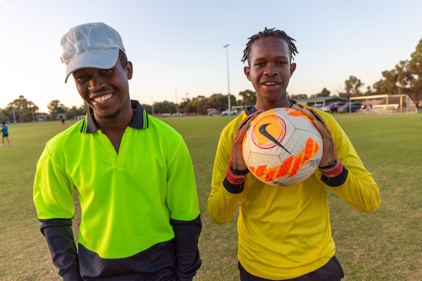 Zwei junge afrikanisch-australische Brüder stehen auf einem Fußballplatz.  Sie lächeln beide und tragen leuchtendes Grün und Gelb.