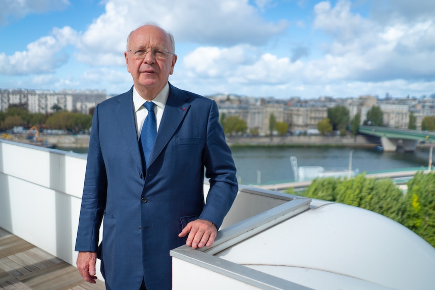 A balding man in a blue suit stands on a rooftop with Paris and the River Seine in the background.