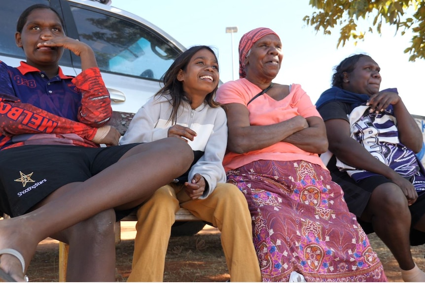 A group of Indigenous people smile and watch football at a country oval