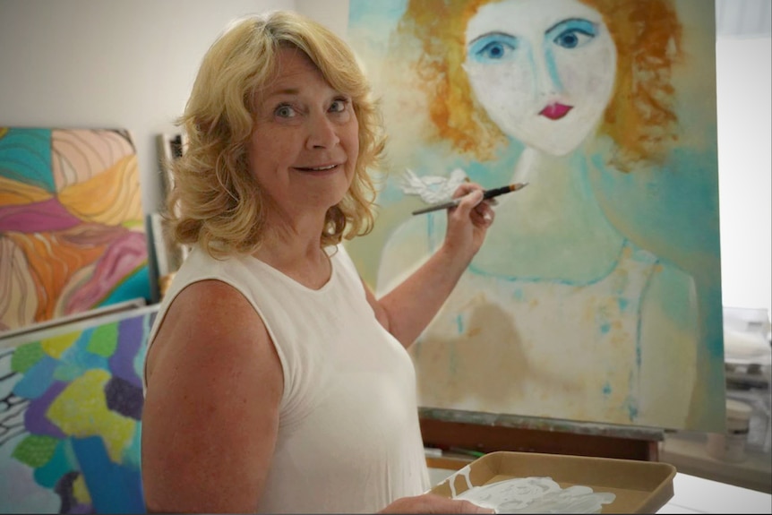 A smiling blonde woman turns to face the camera as she paints a picture of a woman on an easel.