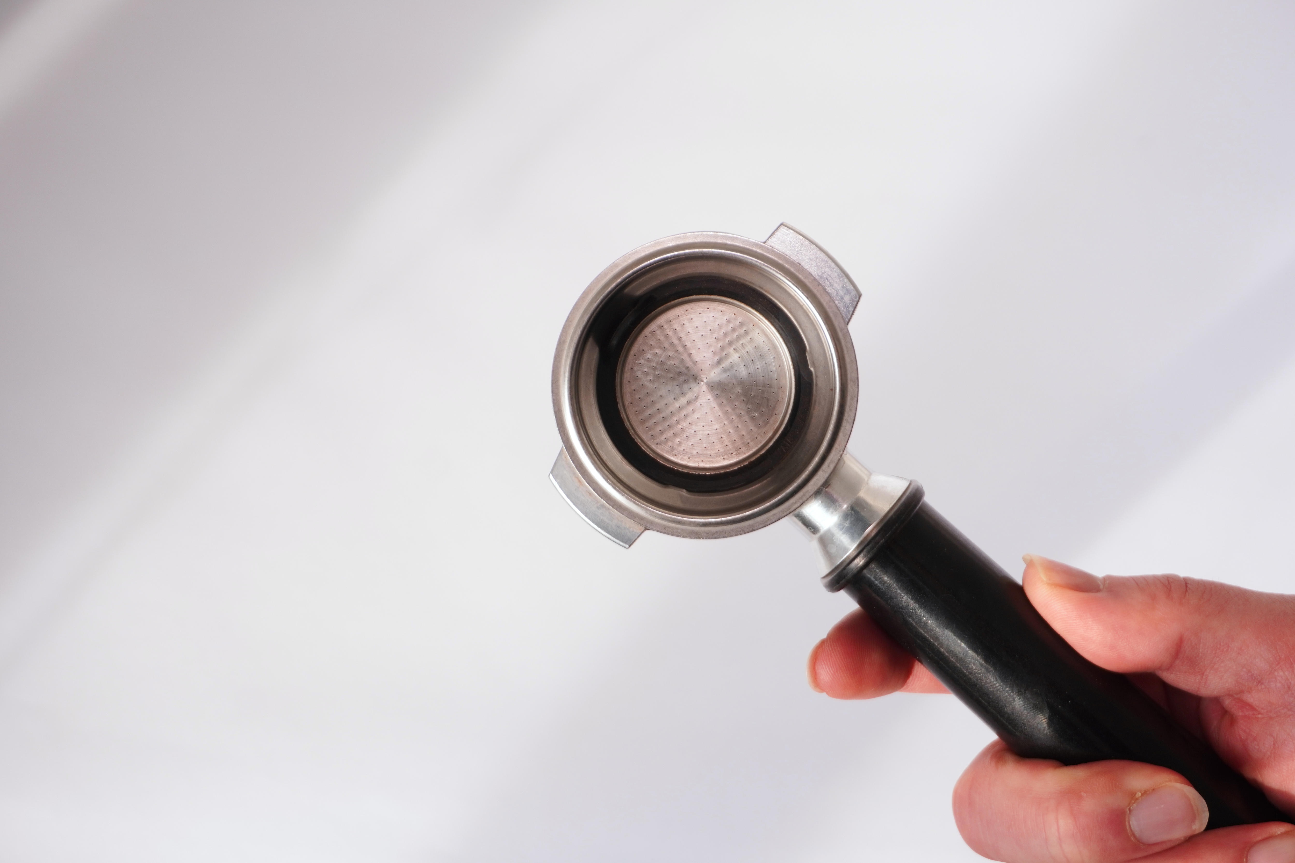 중립적인 배경을 배경으로, 검은색 손잡이가 달린 포타필터를 든 사람이 내부의 정교한 메쉬 필터를 과시하고 있습니다.