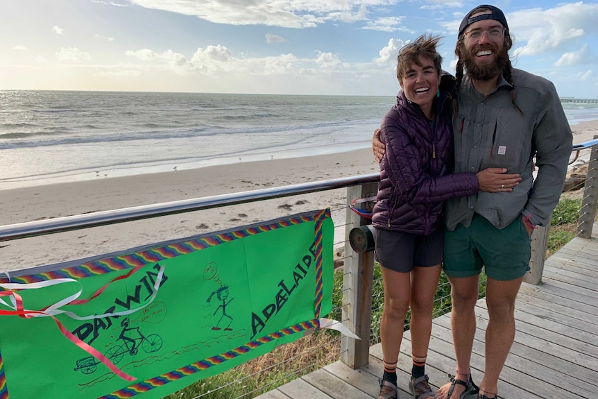 A woman and man wear big smiles as they stand near a green sign that says "Darwin to Adelaide". The ocean is the backdrop.