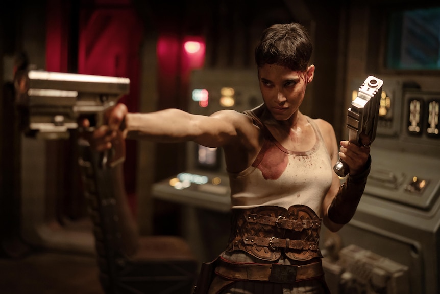 A film still of Sofia Boutella in a bloody dirty top, holding a gun in each hand, one pointing at the screen