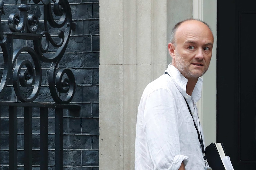 A man with his sleeves rolled to his elbows looks back as he walks towards a door