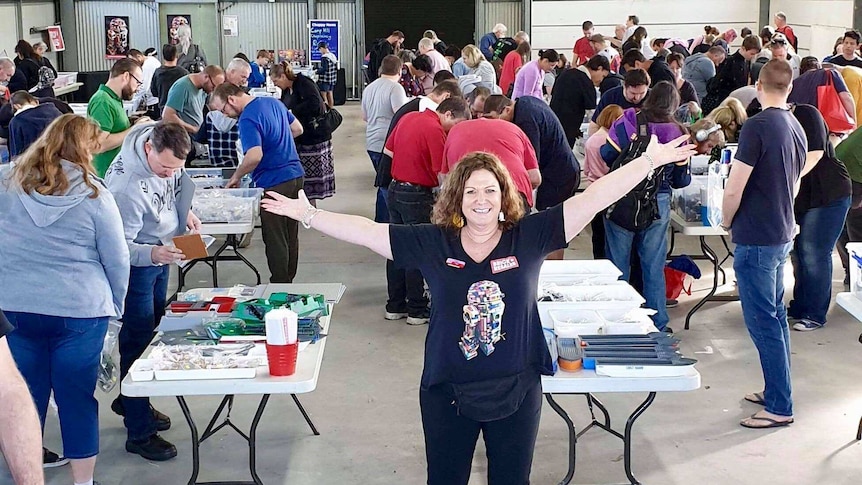 Woman stands with arms outstretched in front of dozens of people poring over boxes of Lego parts.