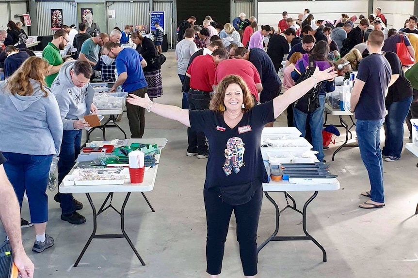Woman stands with arms outstretched in front of dozens of people poring over boxes of Lego parts.
