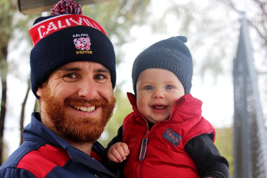 A man dressed in blue and white club supporter clothes smiles at the camera while holding a baby