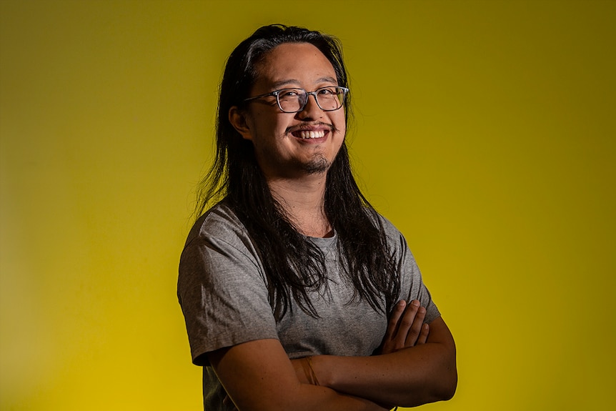 Colour photo of artist Jason Phu smiling with arms crossed and standing in front of a yellow coloured wall.