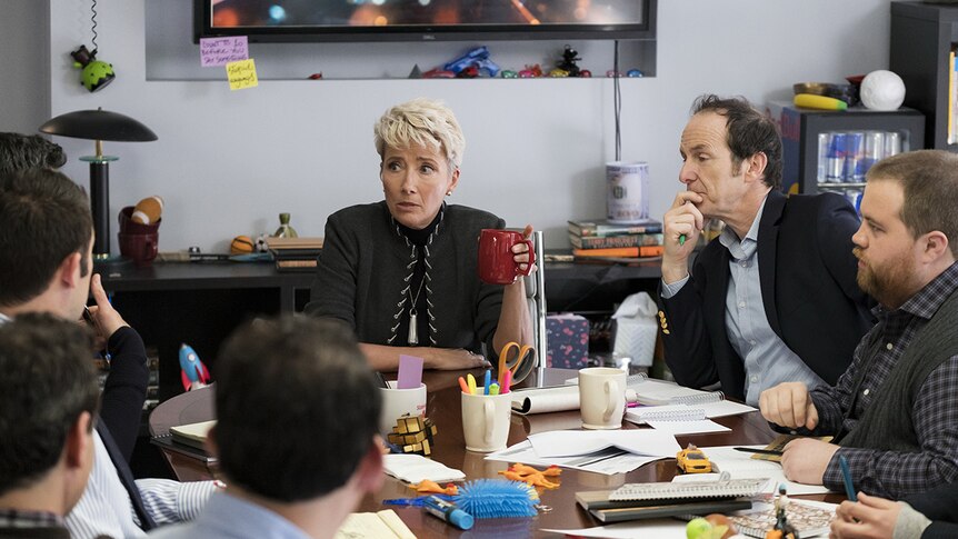 A woman sits at messy meeting table with a group of men in an office space.