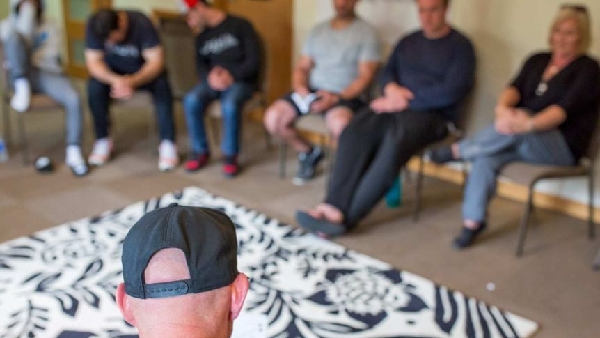 In the foreground is the back of a head, in the background  blurred people sitting in seats at rehab.