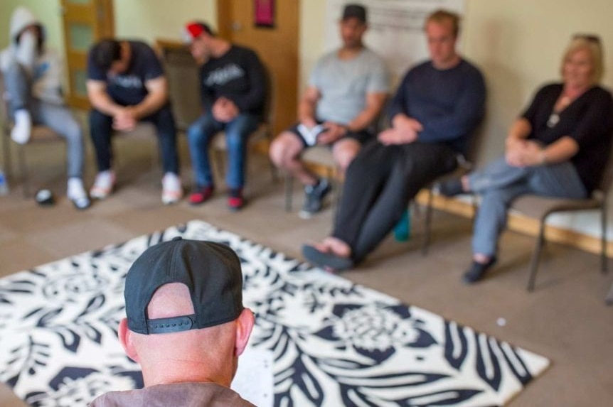 In the foreground is the back of a head, in the background  blurred people sitting in seats at rehab